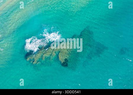Vue de haut en bas d'une formation rocheuse submergée sous une eau turquoise limpide avec de douces vagues se brisant à la surface. La lumière du soleil met en valeur les textures sous-marines, créant une scène côtière vibrante et rafraîchissante. Banque D'Images