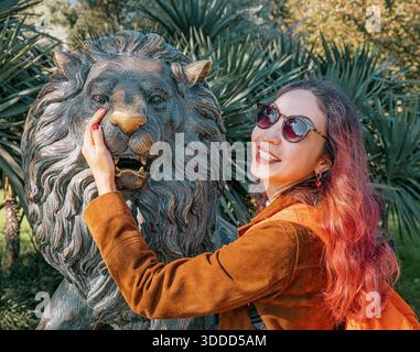 6 novembre 2025, Batoumi, Géorgie : jeune femme souriant, portant des lunettes de soleil, et touchant doucement une grande statue de lion en bronze tout en explorant un parc ensoleillé Banque D'Images
