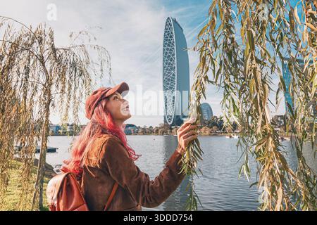 4 novembre 2025, Batoumi, Géorgie : jeune femme souriante, touchant des feuilles de saule avec des gratte-ciel de Batoumi en arrière-plan Banque D'Images