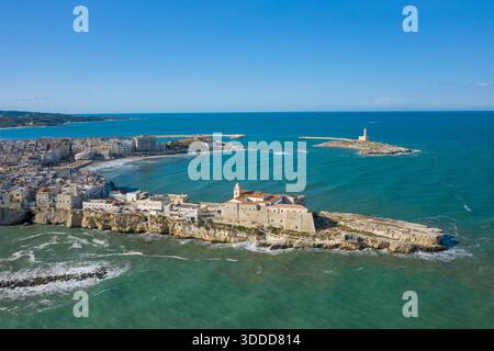 Une vue aérienne panoramique capture la vieille ville historique de Vieste, en Italie, perchée sur des falaises de calcaire escarpées avec le phare de Santa Eufemia debout sur une petite île au large. La mer Adriatique turquoise et le ciel bleu immense encadrent ce paysage côtier méditerranéen emblématique. Banque D'Images