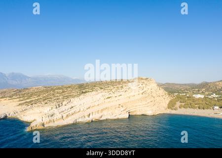 Vue imprenable sur un promontoire de calcaire blanchi par le soleil s'élevant au-dessus de la mer d'un bleu profond à Matala, en Crète. La falaise accidentée rencontre le littoral tranquille sous un ciel clair, avec des montagnes lointaines à l'horizon. Banque D'Images
