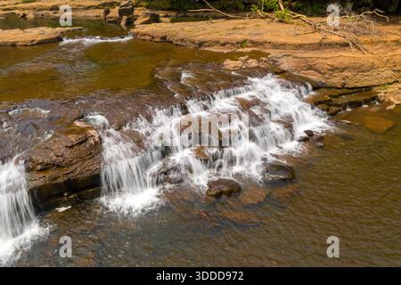 L'eau claire coule doucement sur une série de corniches plates et texturées dans une rivière peu profonde près de Tad Tayicsua dans le sud du Laos. La lumière du soleil met en valeur les tons terreux et les ondulations subtiles, créant une scène tropicale tranquille avec des roches exposées et une végétation minimale. Banque D'Images