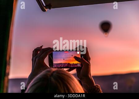 Un bon emplacement dans le ballon à air chaud promet les meilleures vues : une femme filme un ballon à air chaud voisin au lever du soleil à Göreme, Cappadoce, Turquie, avec son smartphone. Les vols commerciaux en ballon au-dessus de la Cappadoce remontent à 1991, fondés par la pilote britannique Kaili Kidner et son mari suédois Lars-Eric Möre ; aujourd'hui, environ 27 entreprises agréées exploitent environ 250 ballons, avec jusqu'à 156 vols et 3 000 à 4 000 passagers par jour. Banque D'Images