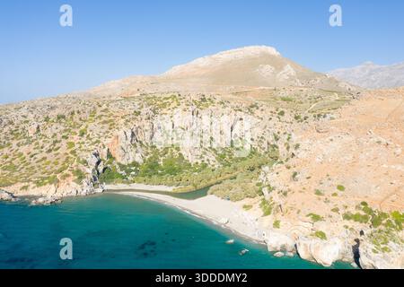 Vue aérienne d'une crique de sable tranquille où une rivière étroite rencontre la mer turquoise, encadrée par des collines rocheuses escarpées et une végétation clairsemée sous un ciel bleu clair en Crète. Banque D'Images