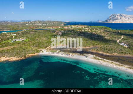 Photographie aérienne de Spiaggia la Salinedda en Sardaigne, avec une baie turquoise vivante, une plage de sable blanc avec parasols, des collines verdoyantes et une montagne rocheuse spectaculaire au loin sous un ciel bleu clair. Banque D'Images