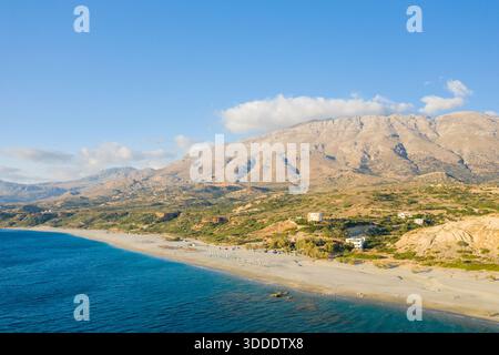 Le rivage sablonneux étendu rencontre la mer bleu profond le long de la côte de Triopetra, en Crète, avec des montagnes escarpées ensoleillées et des maisons dispersées sous un ciel clair. Banque D'Images