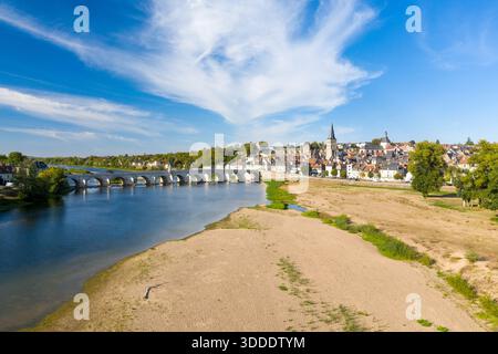 Vue imprenable sur la Charite sur Loire avec son pont de pierre historique traversant la Loire, ses berges sablonneuses au premier plan et la flèche de l'église de la ville s'élevant au-dessus des toits traditionnels. Des nuages tortueux s'étendent sur un ciel bleu vif, soulignant le cadre tranquille au bord de la rivière dans le centre de la France. Banque D'Images