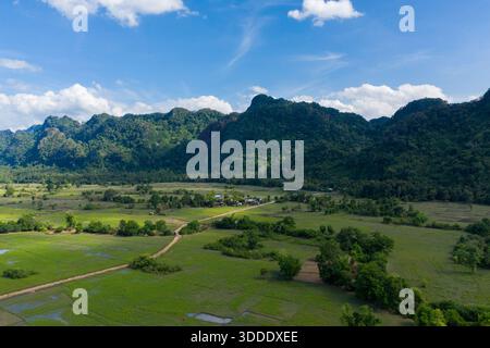 De vastes rizières vertes s'étendent vers des collines calcaires spectaculaires couvertes d'une forêt dense, avec un petit village rural niché à la base. La lumière du soleil éclatante et les nuages dispersés mettent en valeur les textures naturelles et le paysage ouvert de la campagne laotienne. Banque D'Images