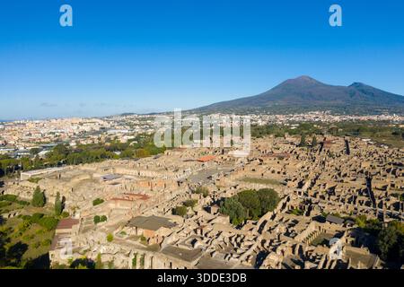 Vue aérienne ensoleillée des ruines antiques de Pompéi avec le mont Vésuve s'élevant au loin sous un ciel bleu vif. La scène capture de vastes vestiges archéologiques, de la verdure éparpillée et le paysage volcanique du sud de l'Italie. Banque D'Images