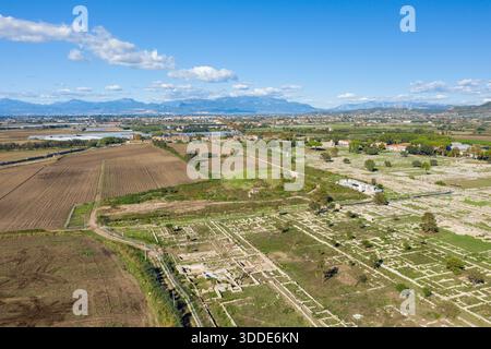 Vue aérienne du site archéologique de Paestum, Italie, avec de vastes fondations en pierre et des ruines situées à côté de champs cultivés. La scène est encadrée par des montagnes lointaines sous un ciel méditerranéen bleu clair avec des nuages éparpillés. Banque D'Images