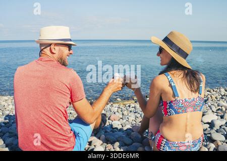 Heureux couple caucasien assis sur la plage de galets avec tasses de dessin animé de café à emporter par la mer noire, concept de vacances, tourisme, détente, style de vie et Banque D'Images