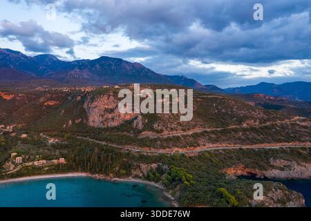 Vue aérienne d'une route sinueuse le long de falaises accidentées au-dessus d'une plage tranquille, avec des maisons dispersées et des montagnes spectaculaires sous un ciel nocturne sombre à Kardamyli. Banque D'Images