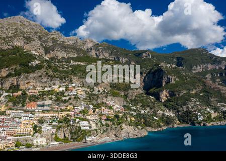 La lumière du soleil illumine les bâtiments colorés à flanc de colline de Positano, en Italie, contre des falaises de calcaire abruptes et un ciel bleu vif. Le paysage emblématique de la côte amalfitaine présente une végétation luxuriante et des eaux méditerranéennes turquoises sous des montagnes spectaculaires. Banque D'Images
