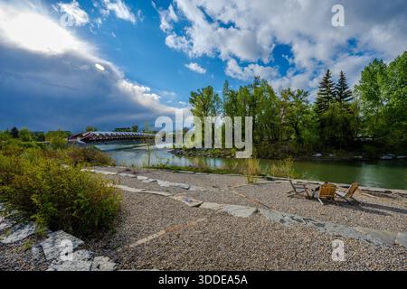 Des chaises en bois se trouvent sur une zone de gravier au bord de la rivière Bow, avec le pont rouge de la paix et des arbres verts sous un ciel partiellement nuageux. Calgary, Canad Banque D'Images