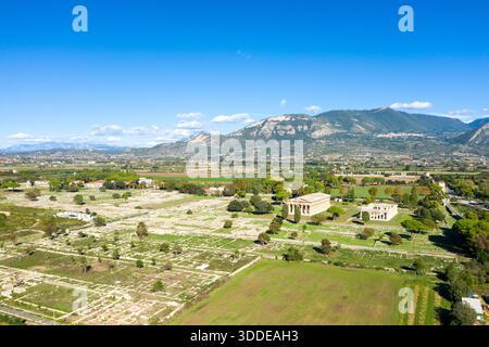 Large panorama aérien de l'ancien site archéologique de Paestum, en Italie, avec de vastes ruines de pierre et deux temples grecs bien conservés au milieu de champs verdoyants. La scène est encadrée par des montagnes lointaines et un ciel méditerranéen bleu vif avec des nuages éparpillés. Banque D'Images
