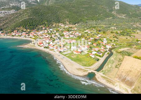 Une vue en grand angle de Kato Vasiliki montre un groupe de maisons au toit rouge le long d'un rivage incurvé, bordé par des eaux turquoises et des collines verdoyantes sous la lumière du soleil. Le paysage comprend un petit canal, des champs ouverts et une atmosphère méditerranéenne tranquille. Banque D'Images