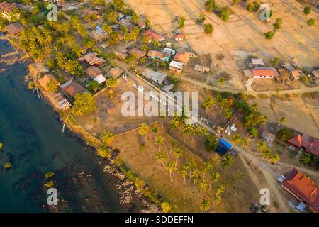 La perspective haute montre un établissement rural le long du fleuve Mékong dans la région des 4000 îles du sud du Laos. Des maisons ensoleillées, des palmiers et des champs dorés de saison sèche créent un paysage tranquille au bord de la rivière avec des bateaux visibles et des motifs de village traditionnels. Banque D'Images