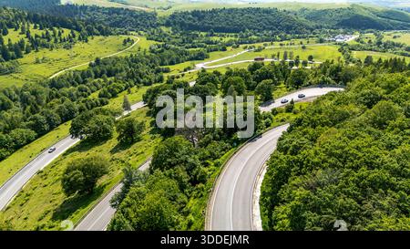 Vue aérienne d'une route de montagne sinueuse traversant le col d'Oituz en Roumanie, entourée de collines boisées verdoyantes et de prairies ouvertes sous un summe partiellement nuageux Banque D'Images