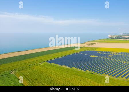 Des rangées de panneaux solaires s'étendent sur des terres agricoles dynamiques près de la côte normande, avec une eau bleue calme et des falaises lointaines sous un ciel d'été lumineux. La scène allie technologie moderne et beauté naturelle du littoral. Banque D'Images