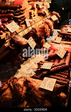 Étal traditionnel de marché avec viandes fumées et saucisses exposées sur des étagères en bois avec des étiquettes de prix manuscrites. Nourriture locale et culture du marché de rue Banque D'Images
