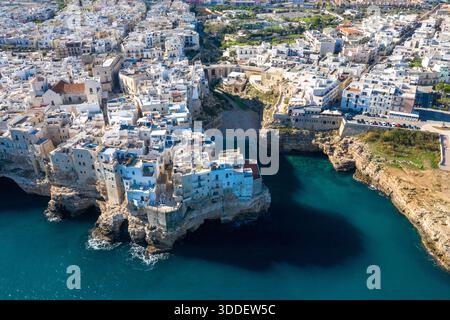 Perspective aérienne expansive de Polignano a Mare dans les Pouilles, en Italie, montrant des bâtiments denses blanchis à la chaux perchés au sommet de falaises de calcaire spectaculaires au-dessus de la mer Adriatique d'un bleu profond. La scène met en évidence le contraste saisissant entre l'architecture historique et la côte accidentée sous la lumière du jour. Banque D'Images