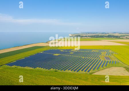 Vue aérienne d'une grande installation de panneaux solaires au milieu de champs jaunes et verts vibrants sur la côte normande, avec un ciel bleu clair et des falaises lointaines créant un paysage frais et durable. Banque D'Images