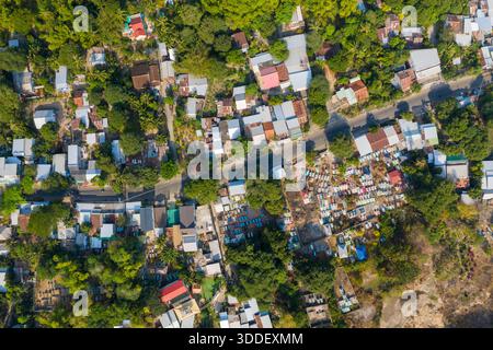 Prise de vue par drone à grand angle d'un petit village vietnamien avec des maisons dispersées, une végétation luxuriante et un cimetière visible. L'image capture la disposition organique et les textures vibrantes de la vie rurale en Asie du Sud-est. Banque D'Images