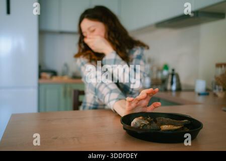Femme éprouvant des nausées et le dégoût d'aliments gâtés, couvrant la bouche et repoussant une assiette de poissons non comestibles, réagissant à des symptômes d'intoxication alimentaire ou de toxicose dans la cuisine Banque D'Images