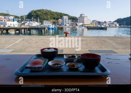 Un plateau avec riz, sashimi, soupe et assortiment de plats d'accompagnement se trouve sur une table en bois surplombant le port calme et les bâtiments de bord de mer de Nachikatsuura par un matin clair. Banque D'Images