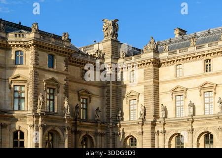Élégante façade en pierre du palais du Louvre à Paris, France, avec des statues détaillées sur le toit et des éléments architecturaux ornés illuminés par la lumière du soleil en fin d'après-midi. Le bâtiment historique se distingue par un ciel bleu vibrant, mettant en valeur son design classique et ses détails sculpturaux. Banque D'Images