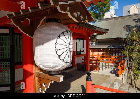 Une lanterne traditionnelle en papier blanc décorée d'un emblème de chrysanthème est accrochée sous des avant-toiTS en bois ornés au sanctuaire Ikuta à Kobe, au Japon. La lumière du soleil met en valeur les faisceaux rouges vifs et les détails architecturaux, créant une scène sereine et accueillante. Banque D'Images