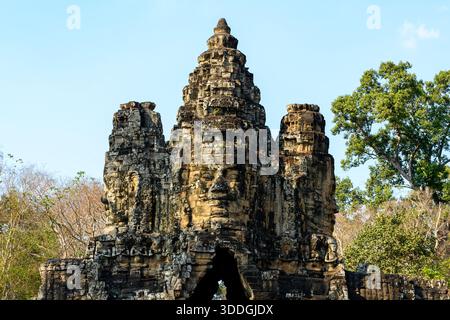Vue frontale d'une porte monumentale en pierre couronnée de visages sculptés sereins à l'entrée d'Angkor Thom près de Siem Reap, Cambodge. L'ancienne architecture khmère s'élève au-dessus des arbres environnants sous la lumière du jour claire, soulignant les détails complexes et les textures altérées. Banque D'Images