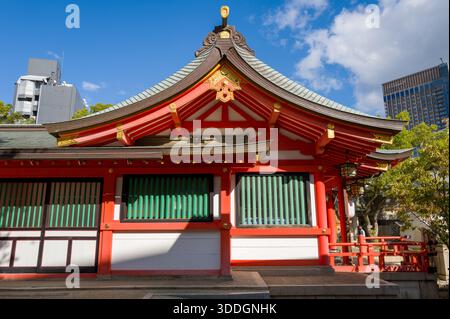 Un pavillon de sanctuaire shinto rouge et blanc vif avec un toit gracieusement incurvé et des accents dorés se dresse sous un ciel bleu au sanctuaire Ikuta à Kobe, au Japon. La lumière du soleil met en valeur les détails architecturaux et les fenêtres en treillis vert, contrastant avec l'horizon urbain en arrière-plan. Banque D'Images