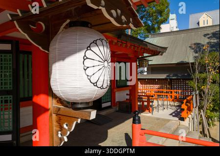 Une lanterne en papier blanc avec une crête de chrysanthème est suspendue sous l'avant-toit d'une structure en bois rouge vif au sanctuaire Ikuta à Kobe, au Japon. La lumière du soleil projette des ombres nettes, soulignant les détails architecturaux et l'atmosphère tranquille. Banque D'Images
