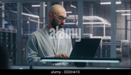 Ingénieur INFORMATIQUE spécialisé utilisant un ordinateur portable dans le hall des serveurs. Man in Light shirt debout et travaillant intentionnellement, l'arrière-plan montre les racks de serveurs Centre de données moderne. Concept diagnostic sur site et maintenance du système. Banque D'Images