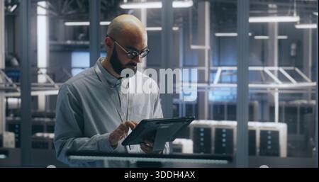 Technicien utilisant une tablette pour la gestion des actifs dans un datacenter. Man Check Assets dans le hall des serveurs. Une collègue marche en arrière-plan. Concept de gestion interactive des actifs et de workflow INFORMATIQUE. Banque D'Images