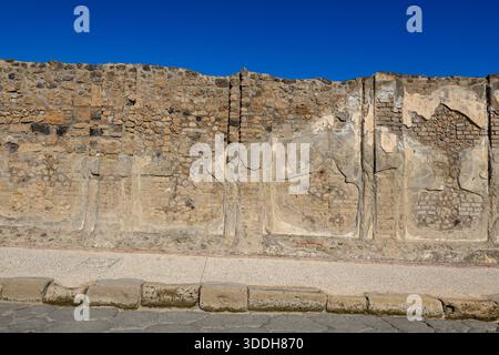 Ancien mur de maçonnerie avec plâtre délavé et briques apparentes ligne une rue à Pompéi sous un ciel bleu vif. L'image met en évidence des textures rugueuses, des détails archéologiques et l'environnement urbain historique de ce site romain. Banque D'Images