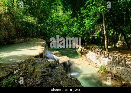 La lumière du soleil illumine un sentier en bois et un mur de pierre longeant l'eau turquoise qui coule sur le calcaire des chutes de Kuang si dans le nord du Laos. La forêt tropicale luxuriante crée un cadre vibrant et serein avec des piscines claires et des textures naturelles. Banque D'Images