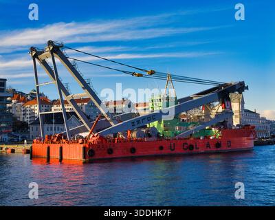 Bergen, Norvège - 13 novembre 2025 : le navire-grue Uglen (IMO 7721079) est ancré sur le front de mer de Bergen, Norvège, encadré par des bâtiments colorés et un ciel bleu vif. Banque D'Images