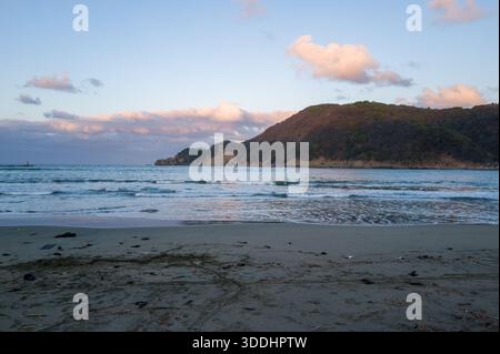 Un littoral de sable calme fait face aux vagues de l'océan, avec une colline couverte d'arbres s'élevant en arrière-plan sous un ciel pastel au crépuscule. La scène capture des textures naturelles, une lumière douce et une ambiance côtière paisible. Banque D'Images