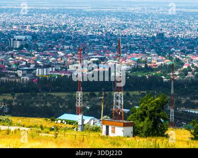 Trois tours de télécommunication s'élèvent au-dessus de petits bâtiments dans un paysage urbain animé de la ville de Bichkek. Les antennes paraboliques et les lignes électriques s'entrecroisent avec des arbres sur une colline herbeuse, créant une vue urbaine étendue. Banque D'Images