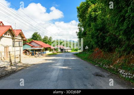 Une route rurale pavée passe devant des maisons avec des toits en métal rouge d'un côté et une végétation verte dense de l'autre, sous un ciel bleu vif avec des nuages dispersés dans les collines du nord du Laos. Les lignes électriques aériennes et un panneau de signalisation sinueux ajoutent des éléments linéaires à la scène tranquille de la campagne. Banque D'Images
