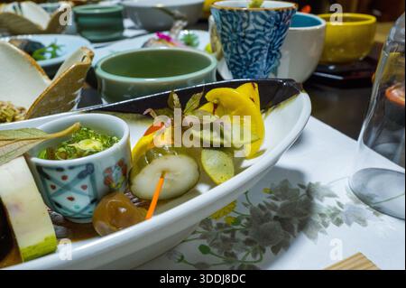 Une sélection vibrante de hors-d'œuvre japonais est habilement agencée dans un plat en céramique, avec des légumes marinés, des garnitures et de petits bols sur une table décorée. La scène est remplie de textures riches, de couleurs fraîches et d'un sens de la présentation culinaire raffinée. Banque D'Images