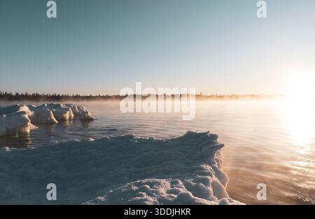 Lever du soleil sur un rivage gelé avec des blocs de glace dispersés et une lumière chaude à travers l'eau. Banque D'Images