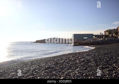 Estran avec des vagues écrasantes sur elle par une journée ensoleillée, avec des bâtiments et une falaise au loin, contreéclairés par le soleil Banque D'Images
