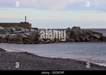 Brise-lames à côté d'une plage par un jour nuageux au coucher du soleil, avec un pêcheur debout sur les rochers Banque D'Images