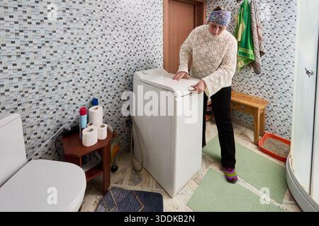 Femme au foyer ferme le couvercle de la machine à laver automatique à chargement par le haut installée dans la salle de bain entre les toilettes et la cabine de douche. Banque D'Images