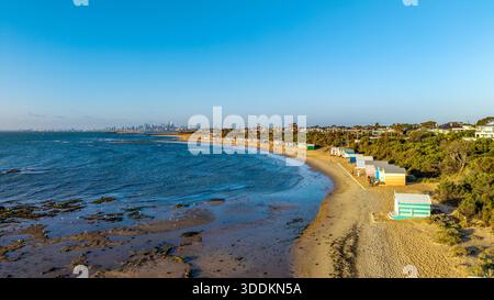 Une vue aérienne met en valeur les emblématiques cabines de bain aux couleurs vives de Brighton Beach et les gens qui se promènent, avec les gratte-ciel de Melbourne à travers Banque D'Images