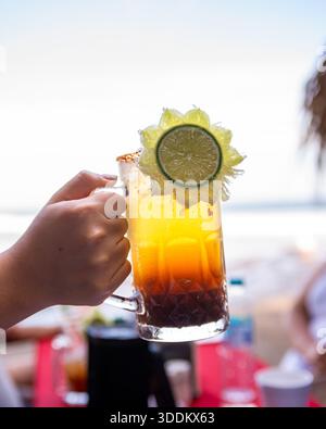 Main féminine tenant une boisson Michelada rafraîchissante et colorée garnie de citron vert. Banque D'Images