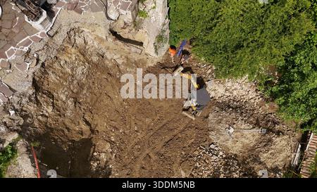 Mini-pelle hydraulique et travailleur dégageant la végétation envahie et les débris sur une surface pavée de la ville près d'une route animée montrant Urban Landscaping Maintenan Banque D'Images